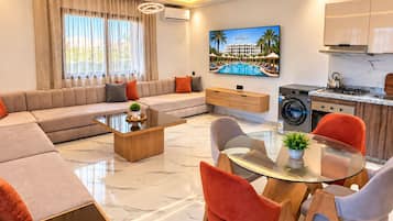Una sala moderna con sofá, mesa de centro y sillas, un área de cocina con estufa y lavadora, y un televisor que muestra un resort.