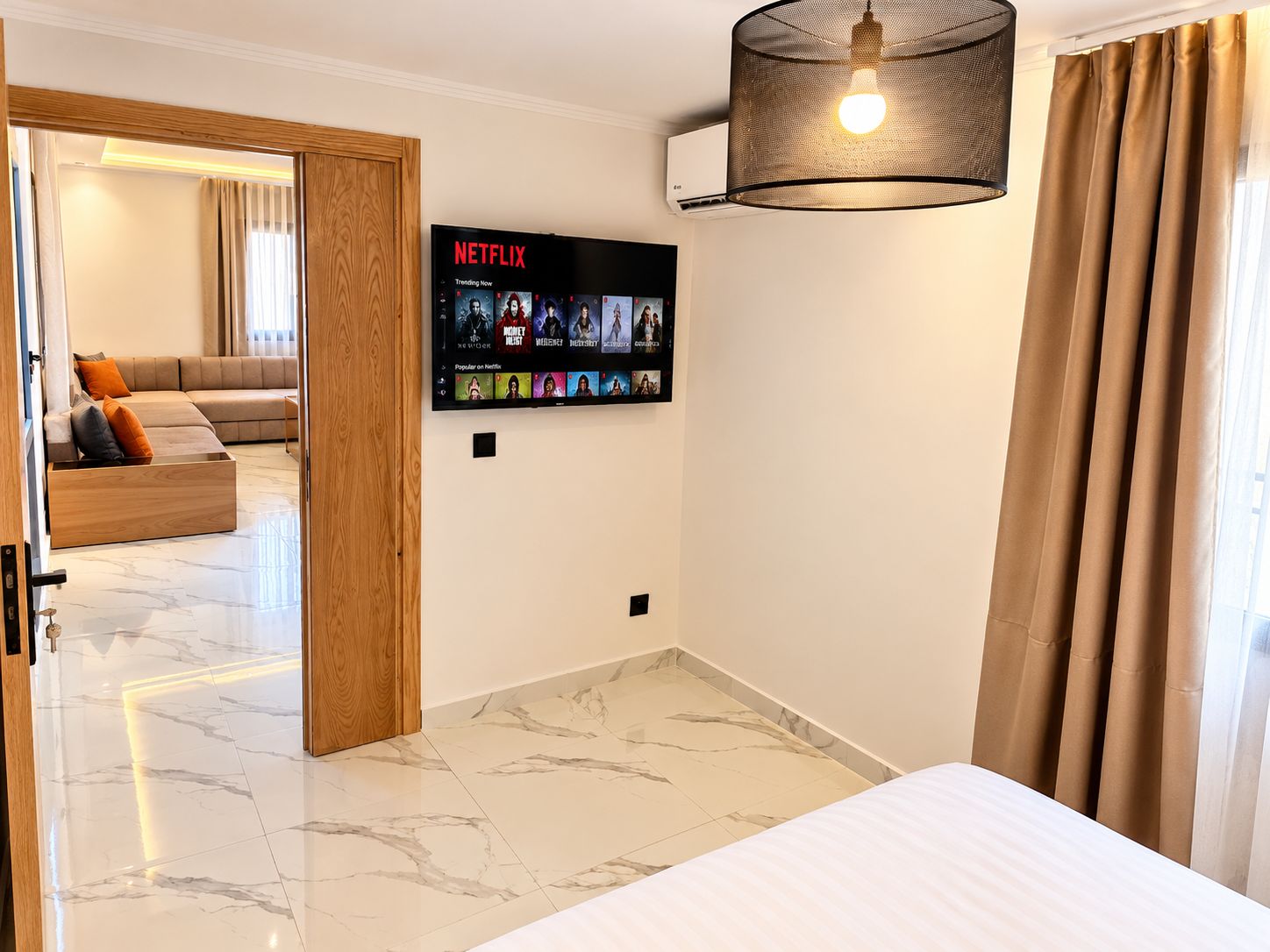A hotel room with a bed, a wall-mounted TV displaying Netflix content, a ceiling light fixture, and a view of a living area with a sofa and curtains.