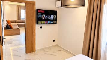 A hotel room with a bed, a wall-mounted TV displaying Netflix content, a ceiling light fixture, and a view of a living area with a sofa and curtains.