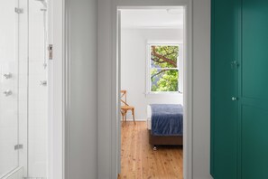 Un couloir étroit mène à une chambre dotée d’une fenêtre, d’un plancher en bois et d’un lit recouvert d’une couverture bleue.