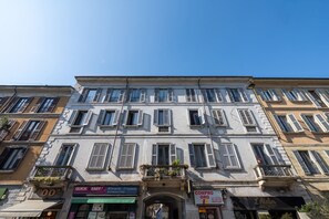 Um edifício com vários andares, com fileira de janelas e varandas, lojas com sinalização e um céu azul límpido.