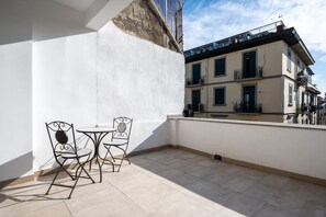 Ein Balkon mit Tisch und zwei Stühlen, eine weiße Wand und ein Gebäude mit Balkonen im Hintergrund.