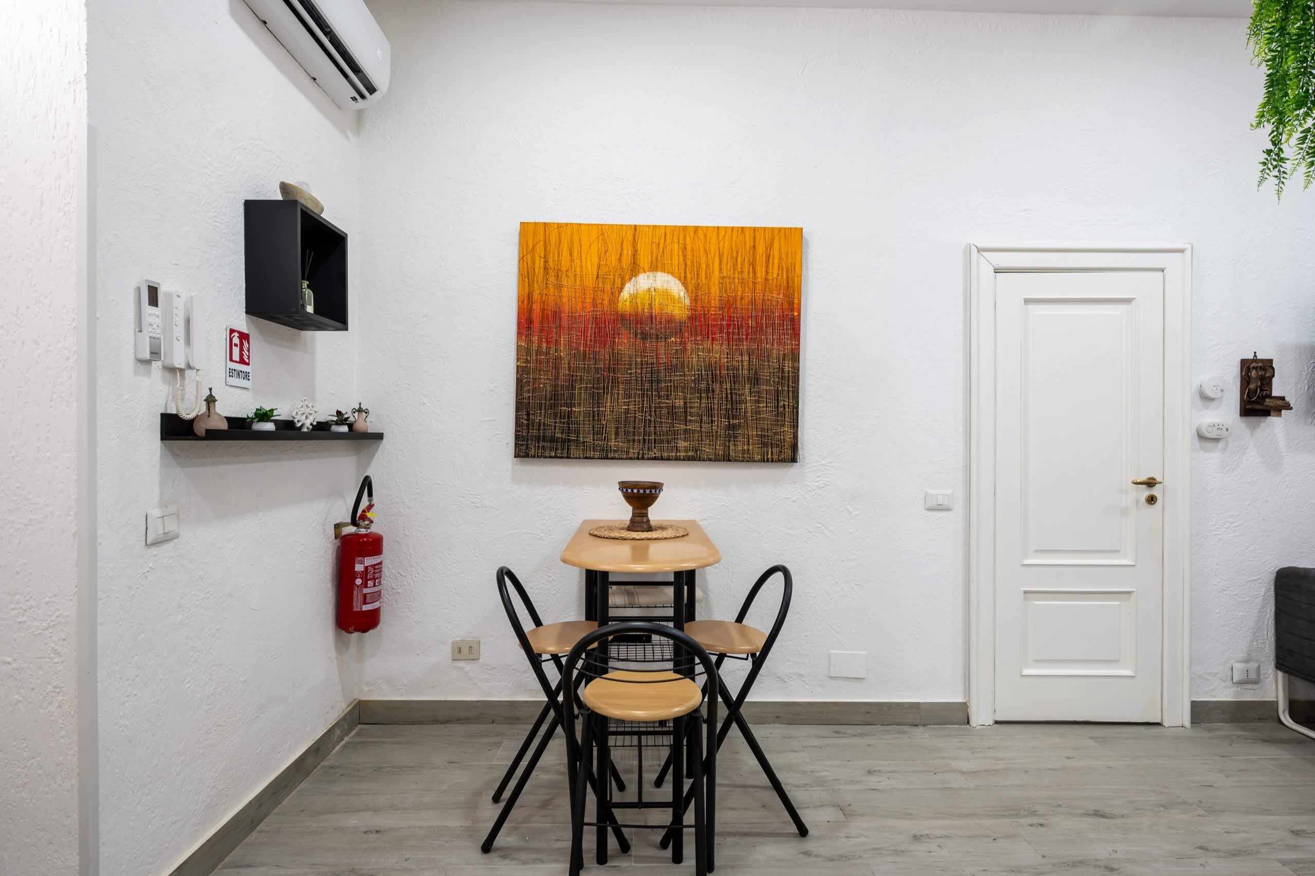 A room with a white door, a table with two chairs, a fire extinguisher, a mounted television, and a painting of a sunset.