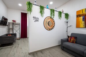A modern living room with a grey sofa, a red curtain, a wall-mounted artwork, and a ceiling-mounted shelf with plants and bottles. - 10 min dall'aeroporto - navetta - self check-in (Naples)