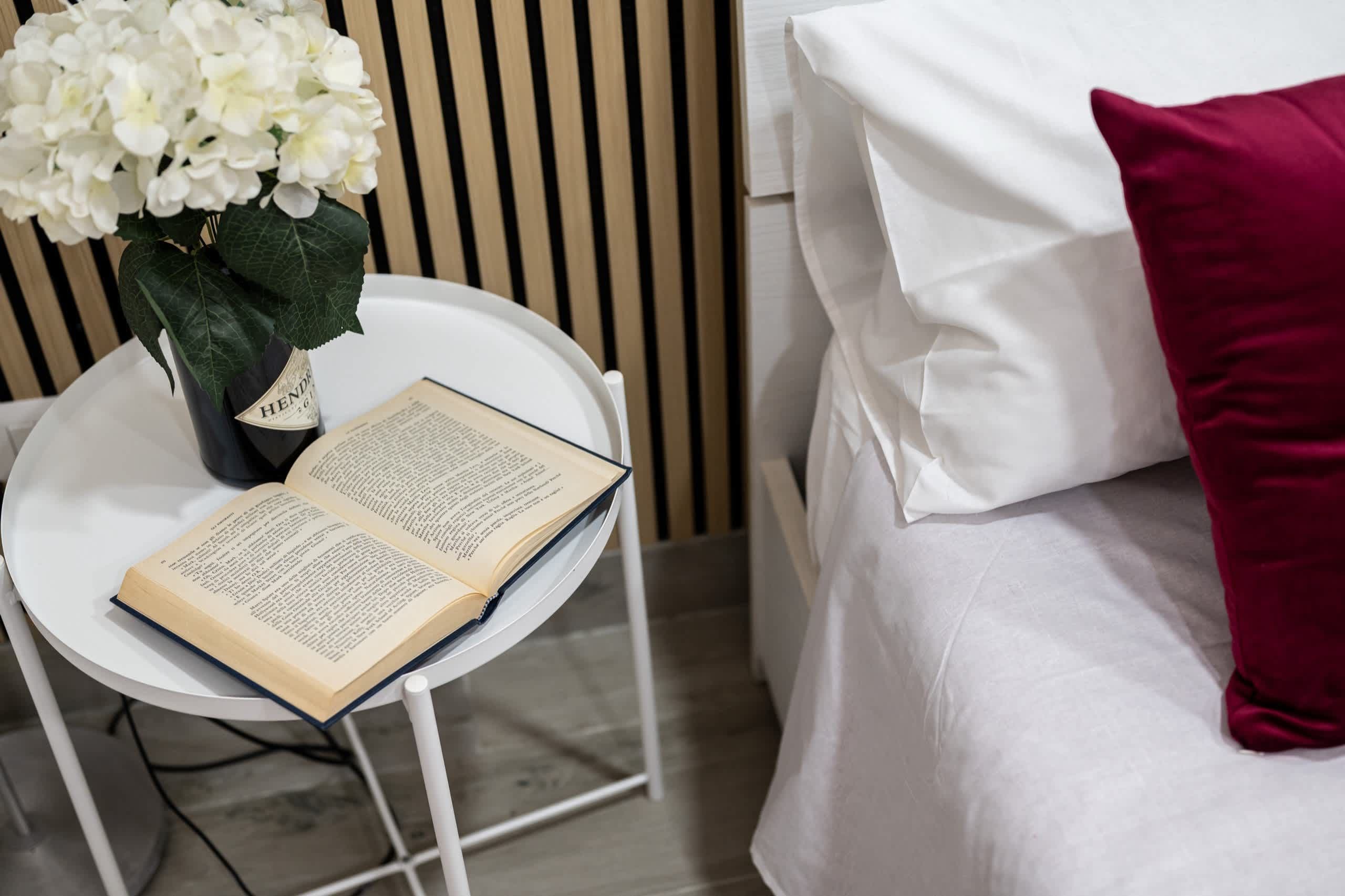 A white bed with a red pillow, a white bedside table with a book and a vase of white flowers.
