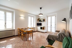 Ein helles Wohnzimmer mit Esstisch, Stühlen, einer Couch, einem Fernseher und einer Lampe.