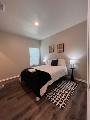 A bedroom with a bed, bedside lamps, a window with blinds, and a patterned rug. - Spacious 2BR Cottage - Relax & Feel at Home (Hurst)