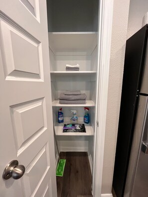 A narrow walk-in closet with white shelves, a folded towel, and cleaning supplies. - Spacious 2BR Cottage - Relax & Feel at Home (Hurst)
