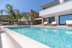 A modern outdoor swimming pool with lounge chairs and a stone building in the background. - Three Bedroom Villa in Nafplio (Nafplio)