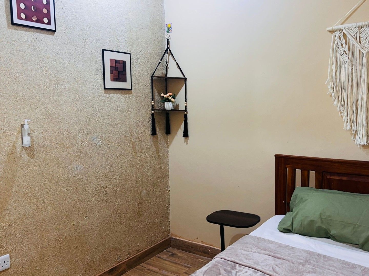 A bedroom with a wooden floor, a bed with a green pillow, a small black table, a wall-mounted shelf with a vase, and a macrame wall hanging.