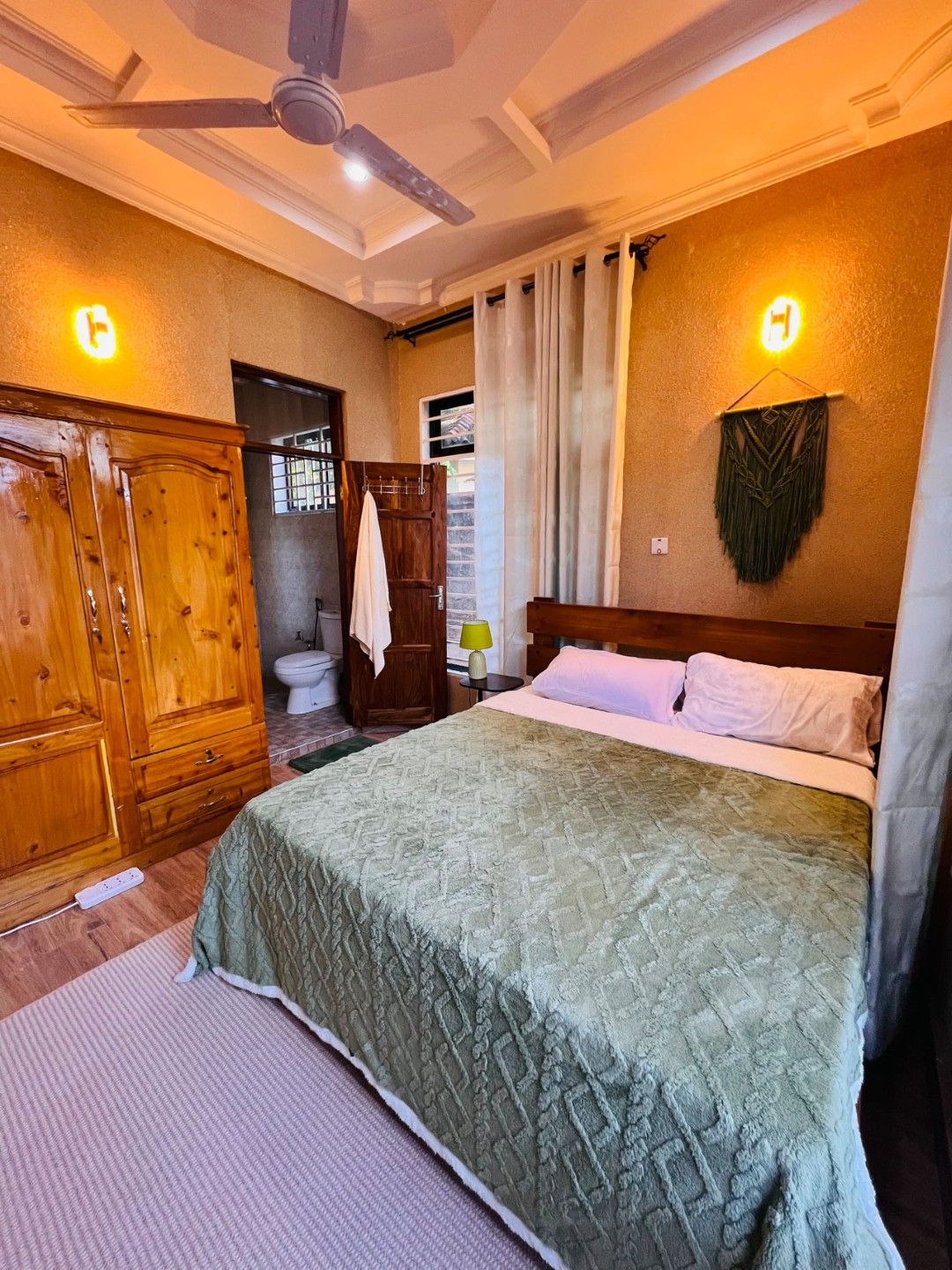 Una habitación de hotel con una cama, armario de madera, baño con inodoro y ventilador de techo.