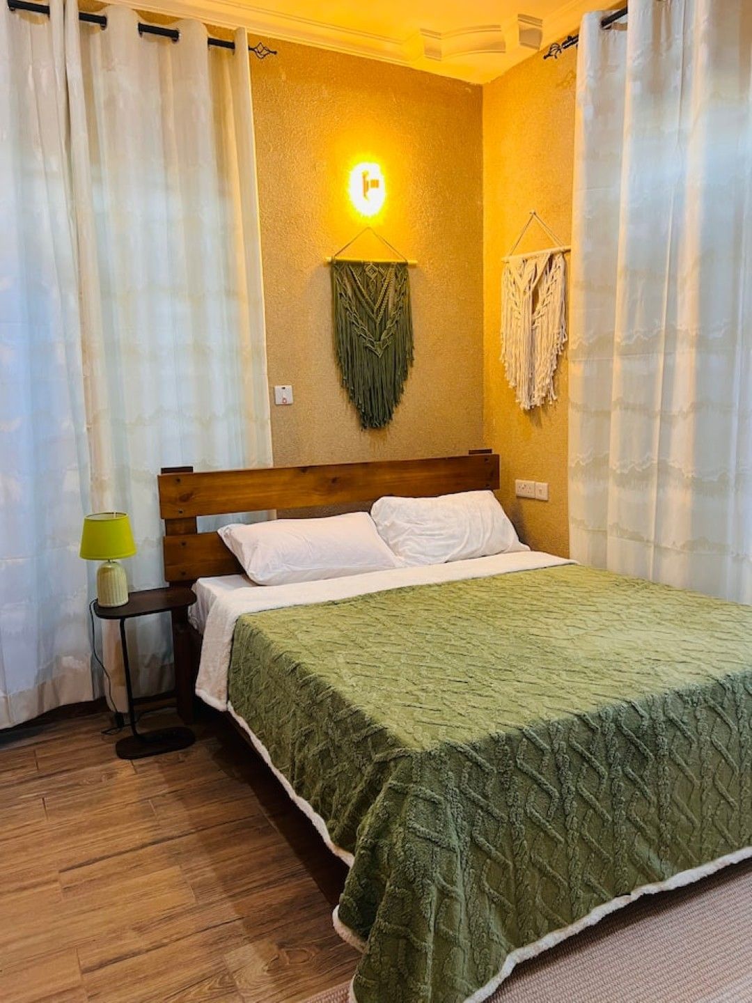 A bedroom with a wooden headboard, a green quilted bedspread, white pillows, a bedside lamp, and a wall-mounted light fixture.