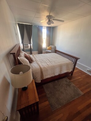 Une chambre à coucher avec un lit, une table de chevet avec une lampe, une chaise, un ventilateur de plafond et une fenêtre avec des rideaux.