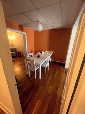 Un coin repas avec une table et des chaises blanches, un ventilateur de plafond et une vue sur une autre pièce où se trouve une lampe.