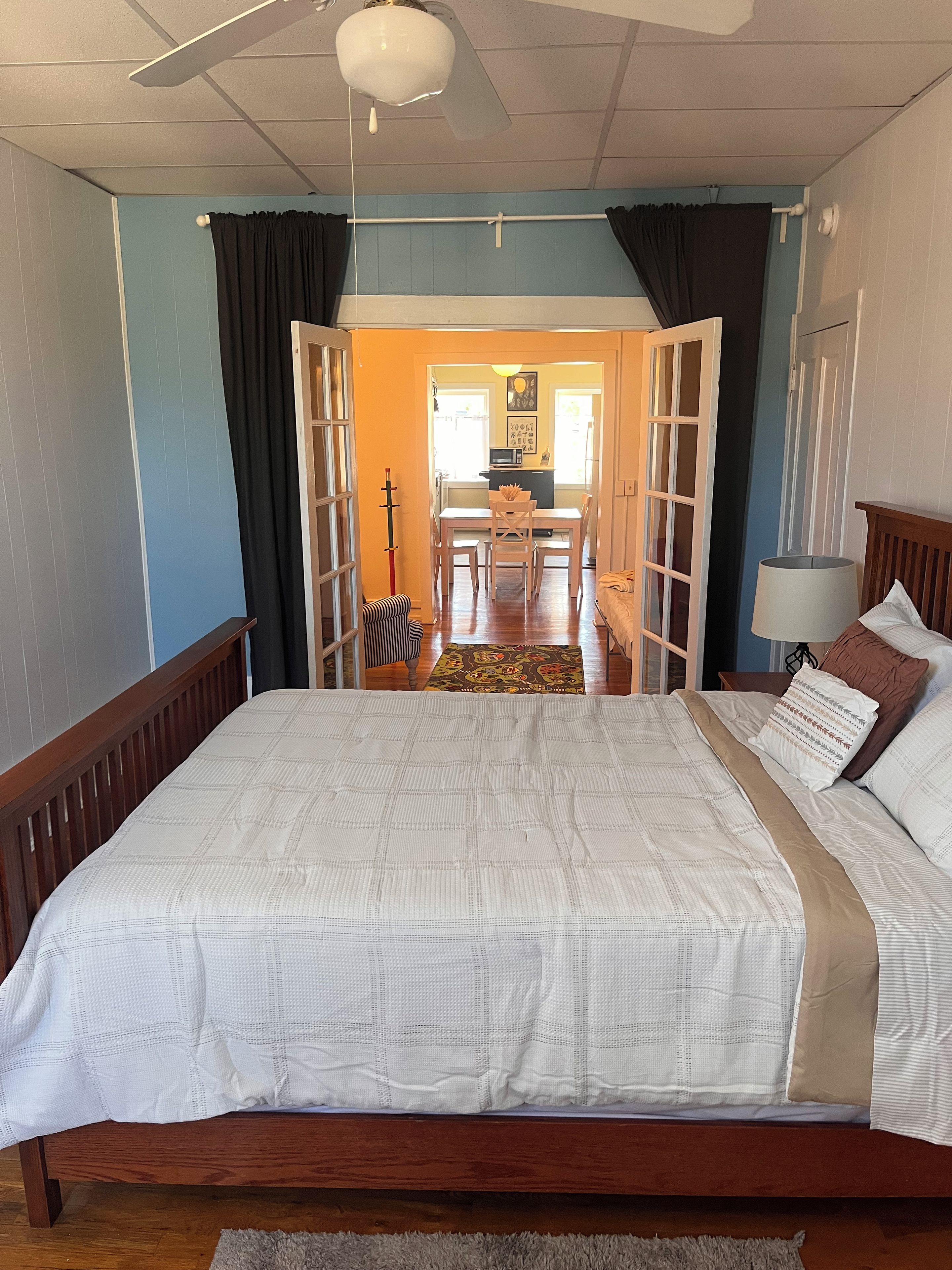 A bedroom with a large bed, a wooden headboard, a nightstand, a lamp, and a view of a dining area through an open door.