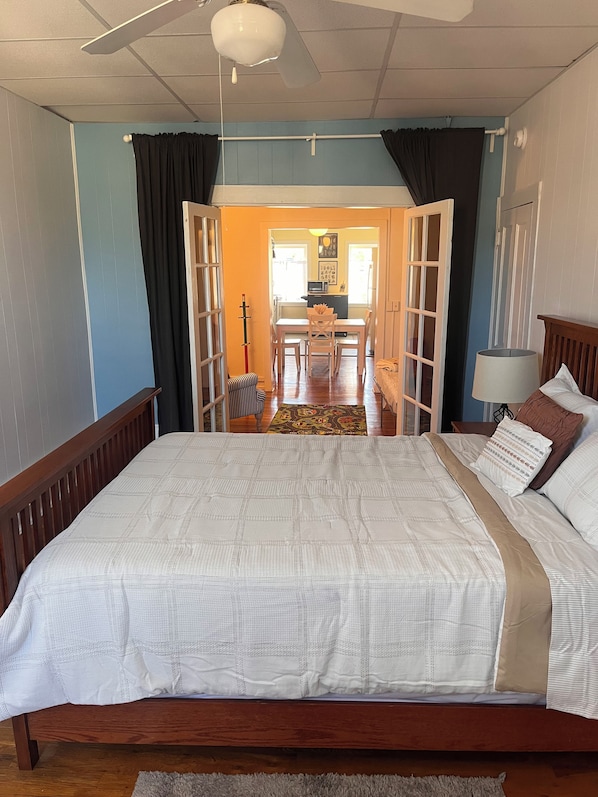 A bedroom with a large bed, a wooden headboard, a nightstand, a lamp, and a view of a dining area through an open door.
