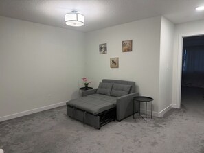 A bedroom with a grey sofa bed, a small round table, and a nightstand with a flower. - Spacious 3BR Duplex Near Edmonton Airport (Edmonton)