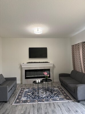 A living room with a fireplace, a flat-screen TV mounted above it, two grey sofas, a patterned rug, and a small table with two cups. - Spacious 3BR Duplex Near Edmonton Airport (Edmonton)