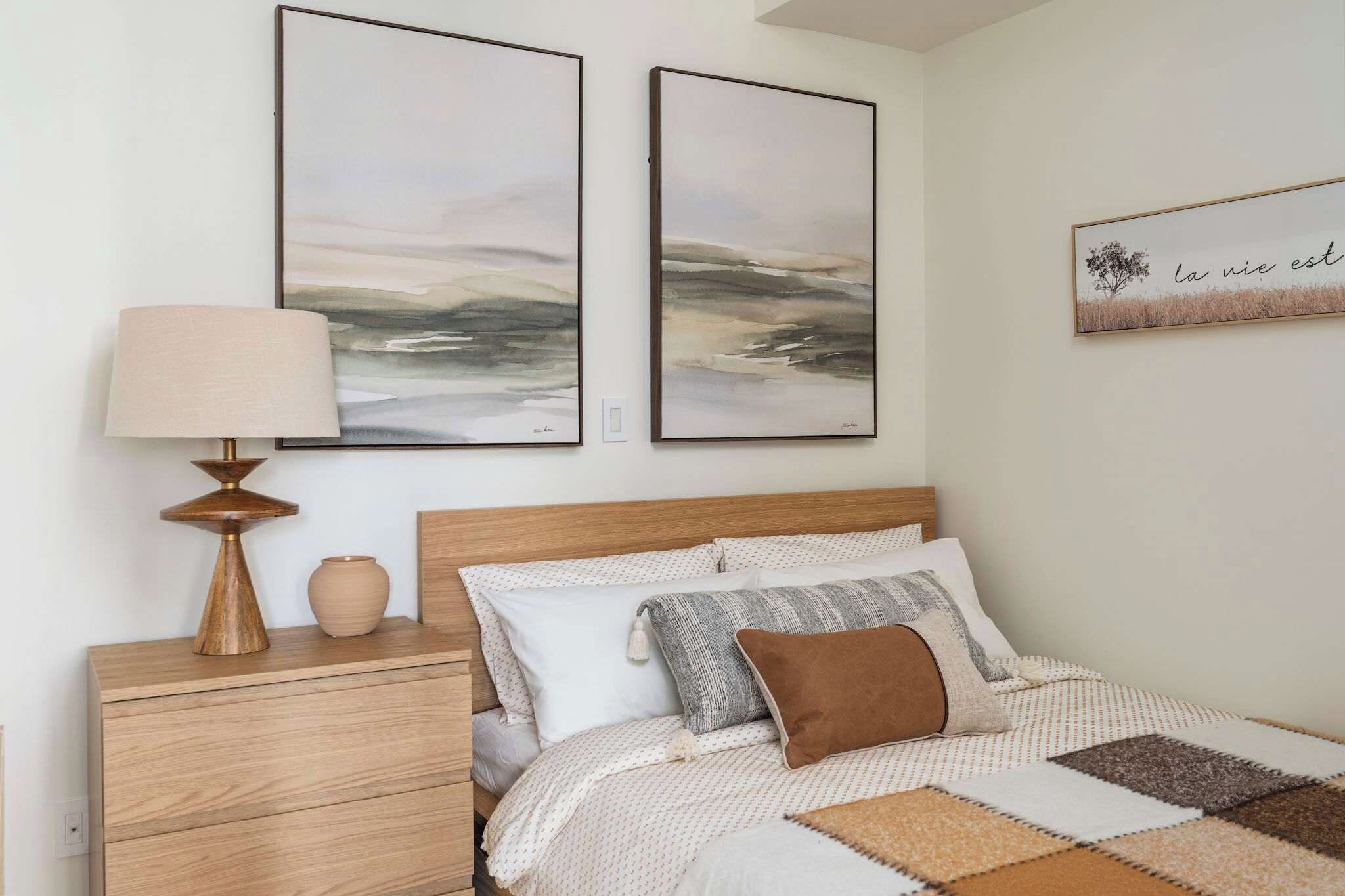 Un dormitorio con cabecera de madera, cama con ropa de cama estampada, mesita de noche con lámpara y dos cuadros abstractos en la pared.