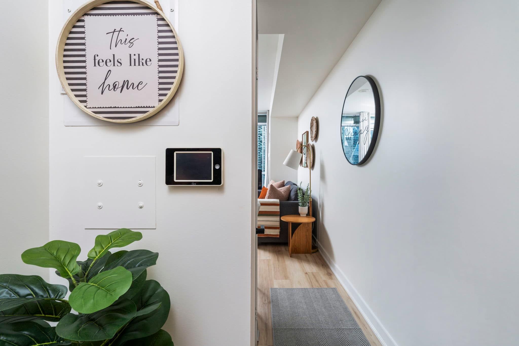 Een hal met een plant, een bord met de tekst 'this feels like home', een wandlamp en een spiegel die op een stadsgezicht reflecteert.