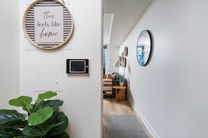 Een hal met een plant, een bord met de tekst 'this feels like home', een wandlamp en een spiegel die op een stadsgezicht reflecteert.