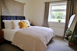 A bedroom with a bed, a nightstand, a window with curtains, and a potted plant. - Kestral Cottage (Brackley)