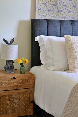 Un lit bien fait, avec une literie blanche et une tête de lit grise, une table de chevet avec une lampe et un vase de fleurs, et un mur orné d’un motif.