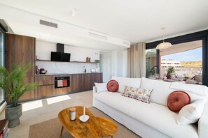 Ein modernes Wohnzimmer mit einem weißen Sofa, einem Holztisch und einer Küchenzeile inklusive Backofen und Mikrowelle.