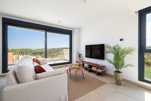 A modern living room with a large window, a flat-screen TV, a sofa, a coffee table, and a potted plant. - Family-friendly 2-bedroom penthouse with a padel court (Mijas)