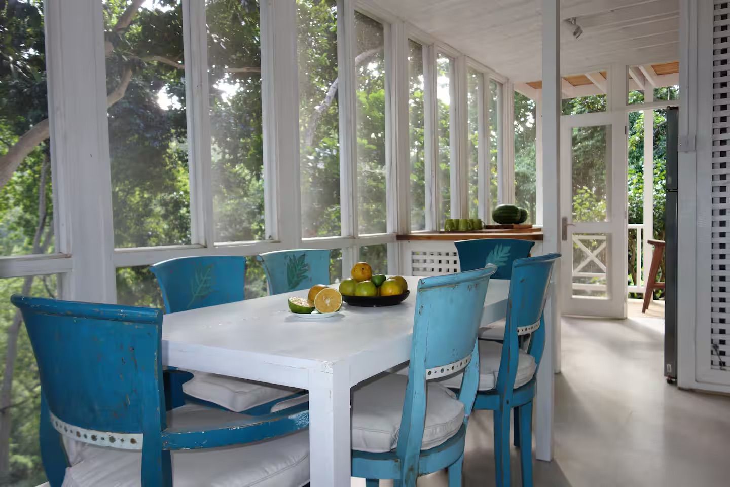 Un espace repas abrité, avec une table blanche et des chaises bleues, un saladier rempli de fruits et une vue sur la verdure environnante.