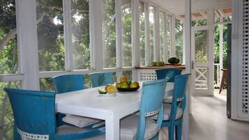 Un espace repas abrité, avec une table blanche et des chaises bleues, un saladier rempli de fruits et une vue sur la verdure environnante.