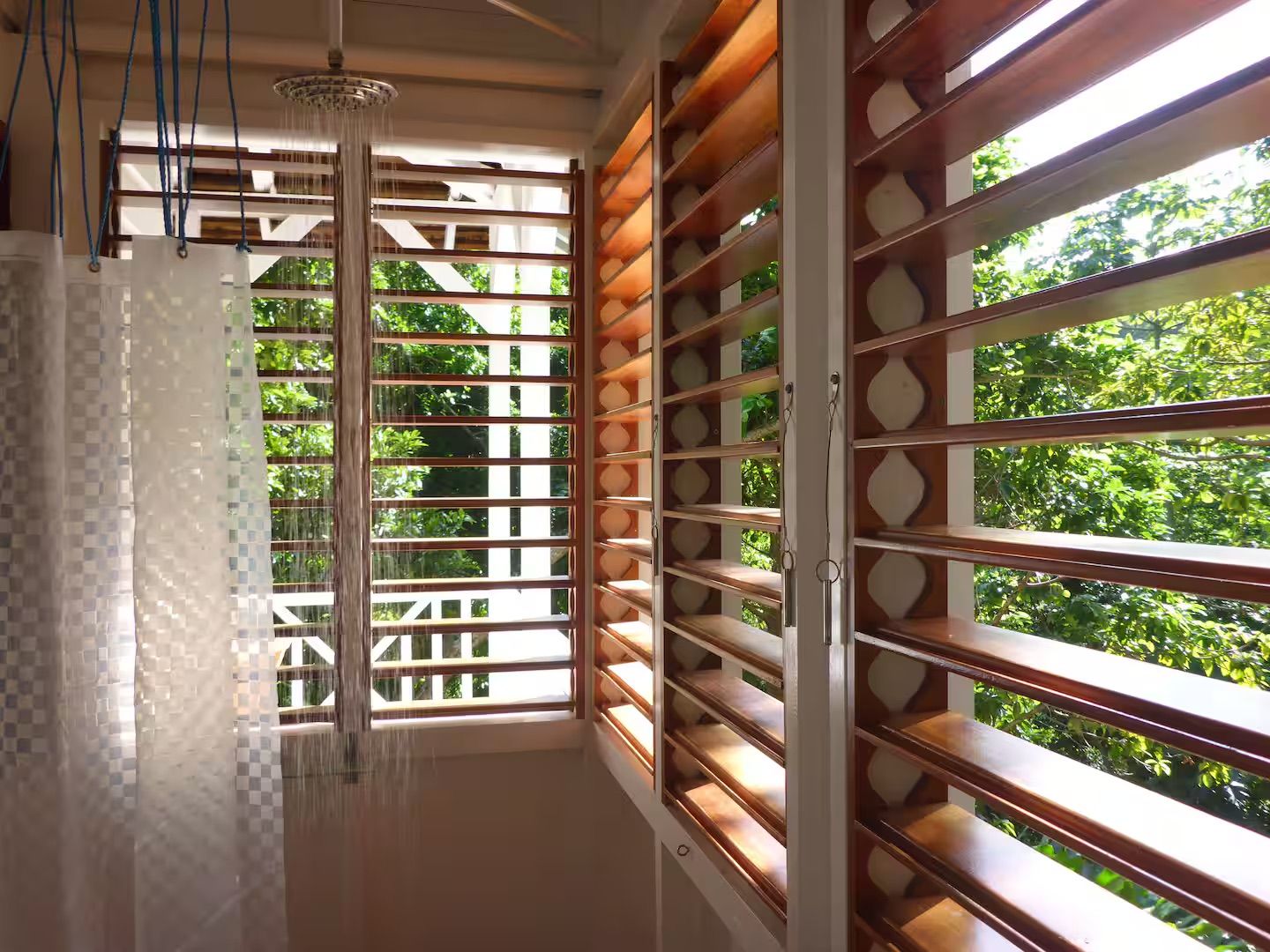 A bathroom with a shower, a window with wooden slats, and a view of greenery outside.