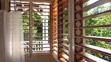 A bathroom with a shower, a window with wooden slats, and a view of greenery outside.