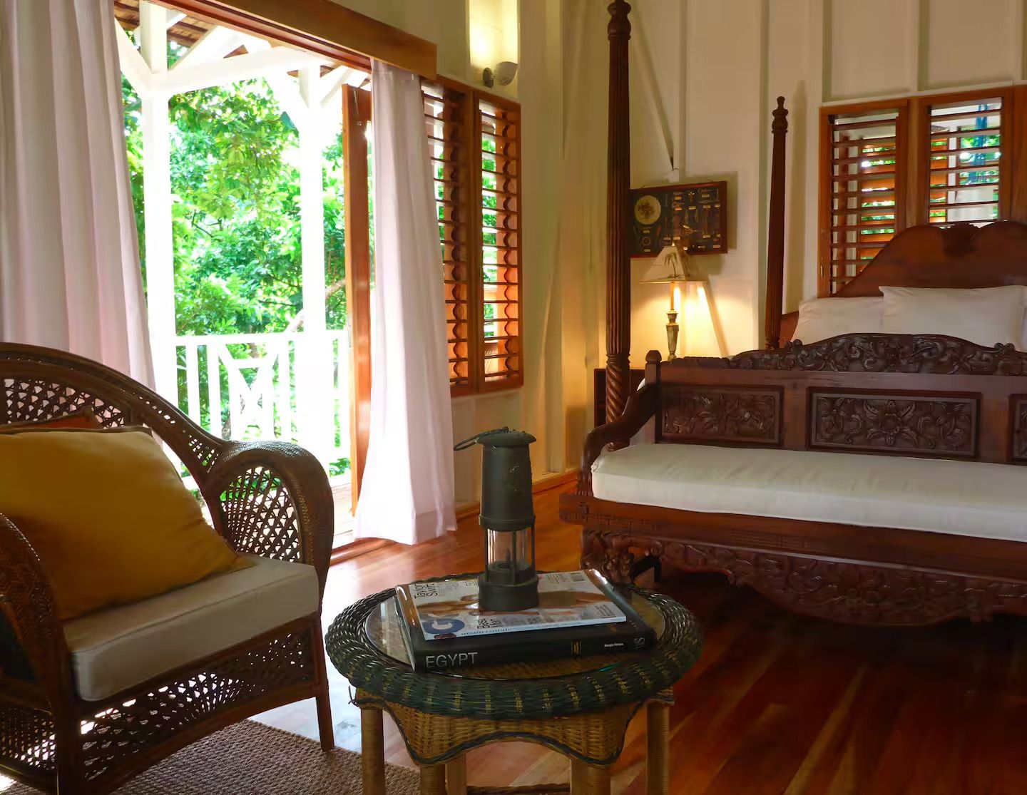A room with a bed, a chair, a table with a book, and a window with a view of greenery.