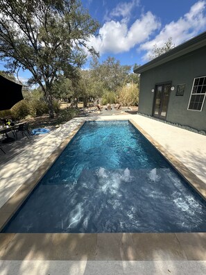 En rektangulär swimmingpool med klart vatten, omgiven av en terrass och en byggnad med en dörr och fönster.