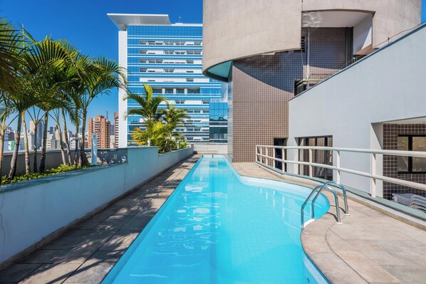 Una piscina de diseño moderno, rodeada por un muro bajo y palmeras. Al fondo, un edificio alto con fachada azul.