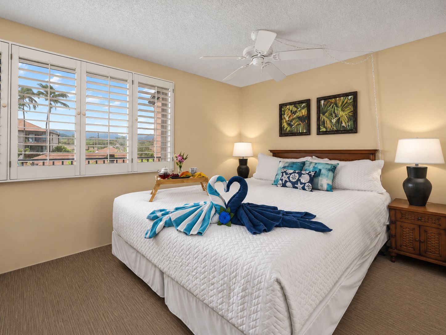 A hotel room with a bed, two bedside lamps, a ceiling fan, a window with blinds, and artwork on the wall.