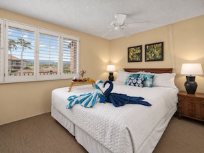 A hotel room with a bed, two bedside lamps, a ceiling fan, a window with blinds, and artwork on the wall.
