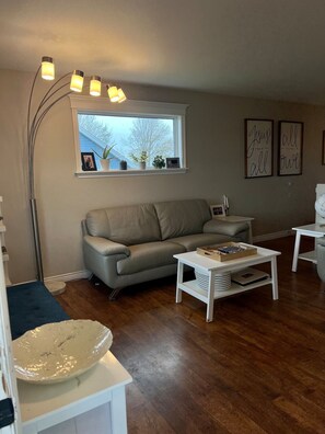 A living room with a sofa, a coffee table, a floor lamp, and a window with plants. - Fenced in pet friendly 3 bedroom home (Stratford)