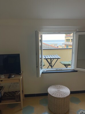 Ein Zimmer mit Fernseher auf einem Schrank, einem kleinen Tisch, einem Korbsessel und einem Fenster mit Blick auf Strand und Gebäude.