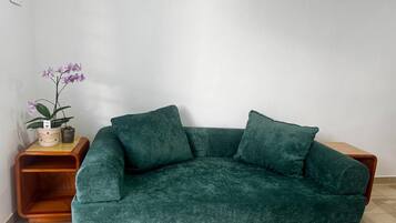 A green velvet sofa with two cushions, a wooden side table with a potted plant, and a white wall.