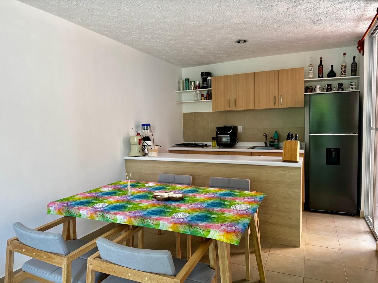A compact kitchen with a dining area, featuring a table with a colorful tablecloth, wooden cabinets, and a stainless steel refrigerator.
