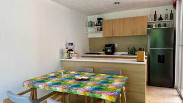 A compact kitchen with a dining area, featuring a table with a colorful tablecloth, wooden cabinets, and a stainless steel refrigerator.