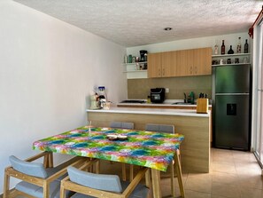 A compact kitchen with a dining area, featuring a table with a colorful tablecloth, wooden cabinets, and a stainless steel refrigerator. - Anáhuac Relaxation Room (Oax.)