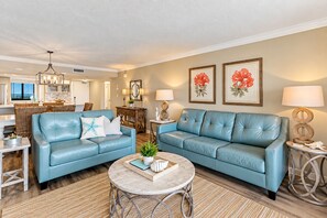 A living room with two blue sofas, a coffee table, and a dining area with wicker furniture. - 5-Star Beachfront Condo, Edgewater Resort #1402 (Miramar Beach)