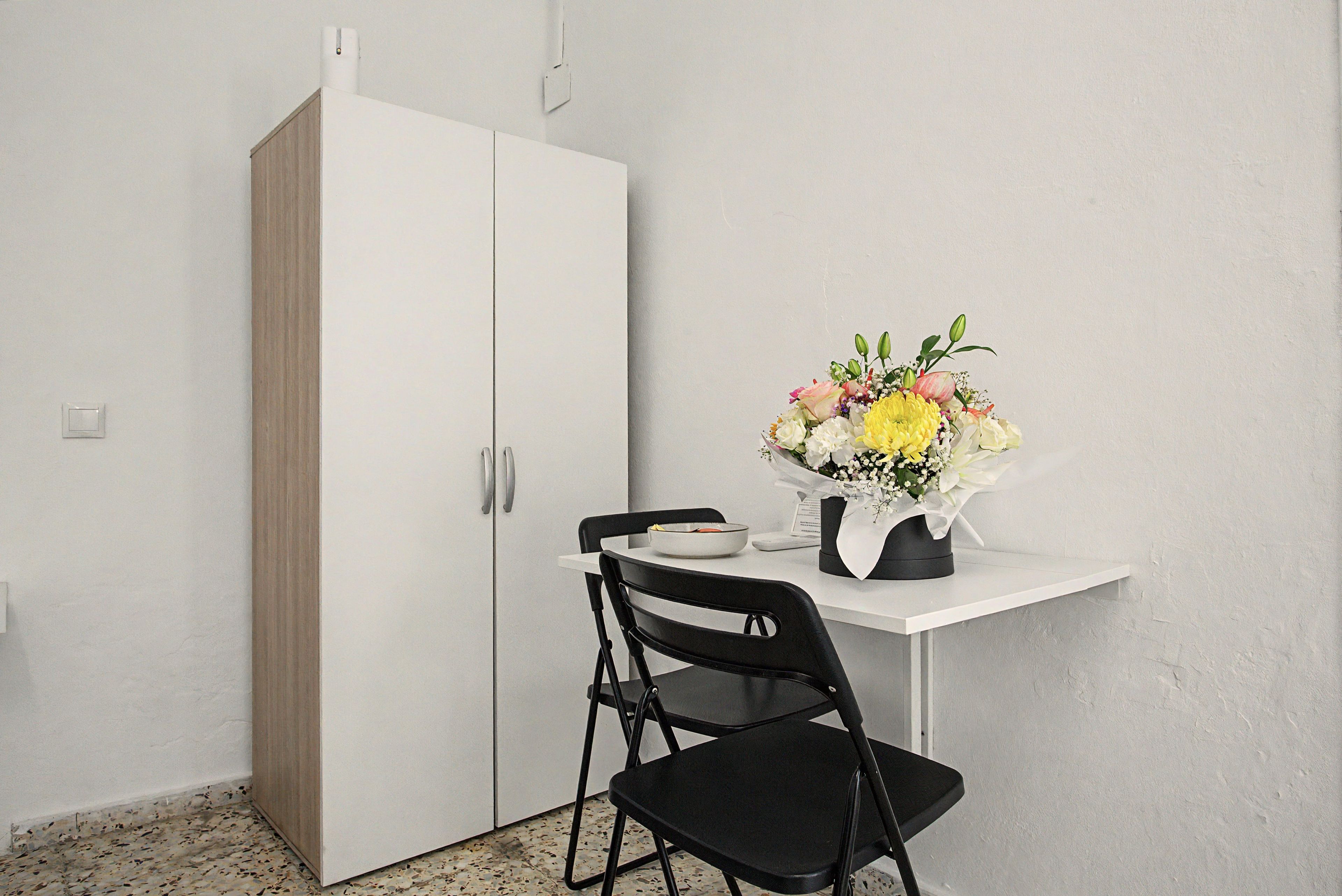 Una kitchenette con un armario blanco, una silla negra y una mesa con un florero.
