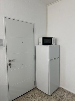 Eine weiße Tür mit Schlüsselloch und einem kleinen Schild, ein weißer Kühlschrank mit einer Mikrowelle darüber und ein Betonboden.