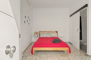 Un dormitori petit i ben organitzat, amb un llit de fusta, un edredó vermell i una petita taula de nit amb un ram de flors.