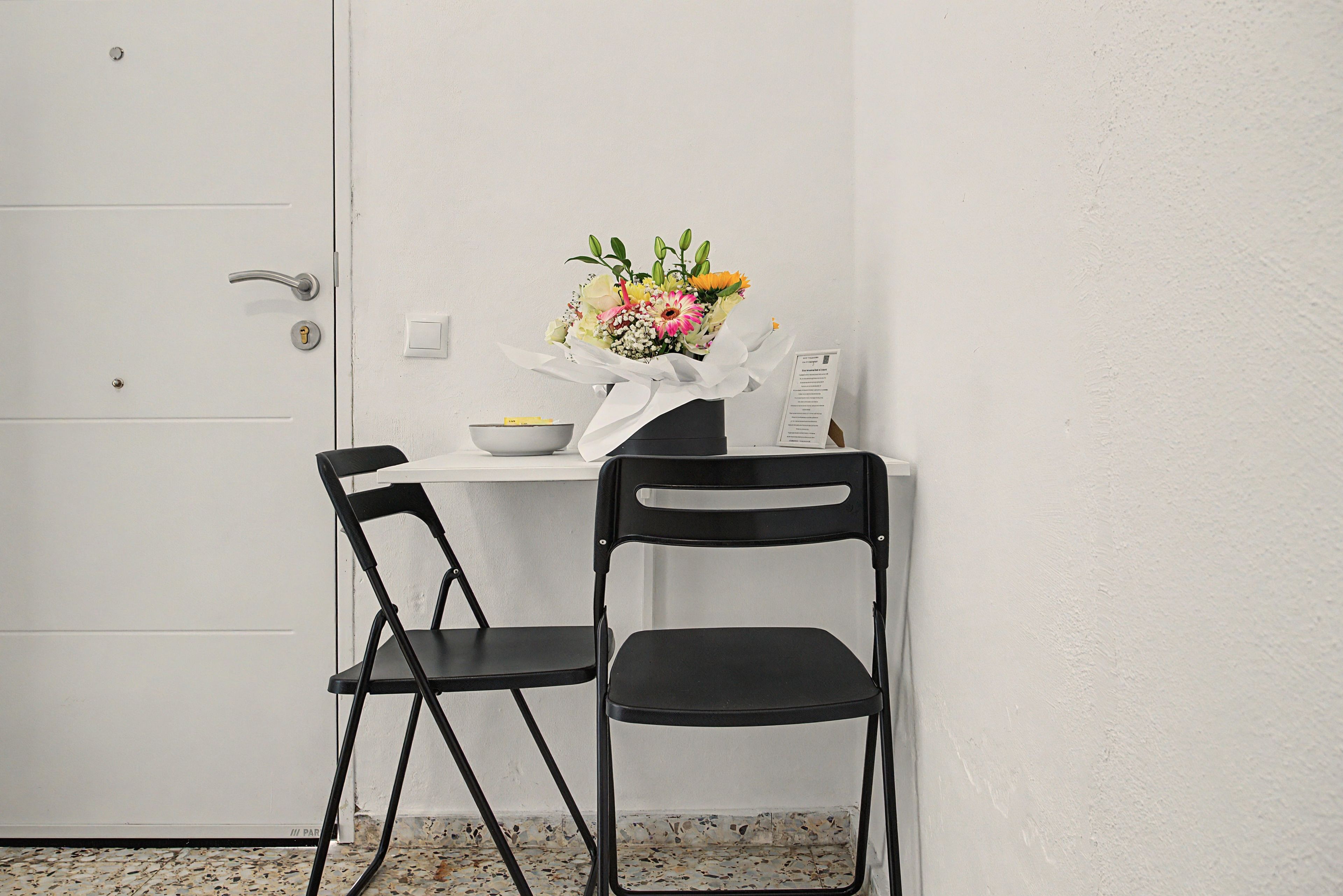 A small room with a white door, a white table with a vase of flowers, two black chairs, and a white wall.