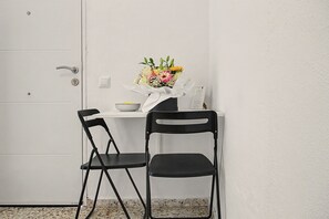 Une petite pièce avec une porte blanche, une table blanche ornée d’un vase de fleurs, deux chaises noires et un mur blanc.